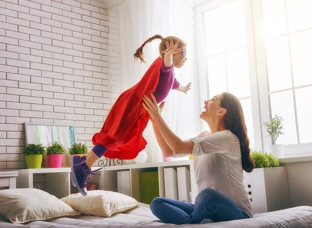 Mother holding up daughter dressed as super hero in bedroom